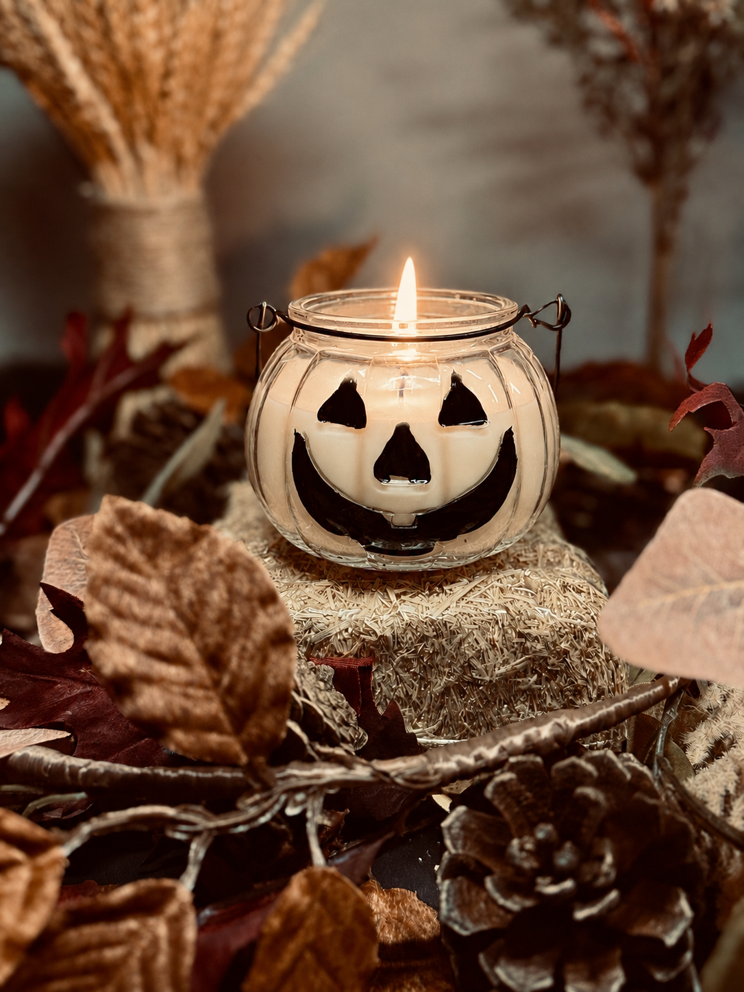 An clear pumpkin-shaped candle with a carved jack-o'-lantern face, lit with a small flame, displayed on a small bundle of hay surrounded by autumn leaves, pinecones, and dried wheat in a rustic setting.