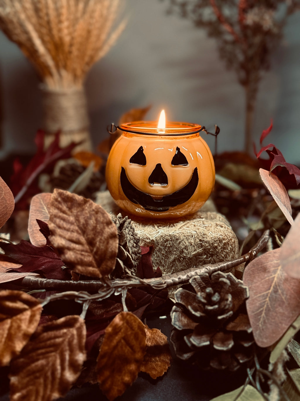 An orange pumpkin-shaped candle with a carved jack-o'-lantern face, lit with a small flame, displayed on a small bundle of hay surrounded by autumn leaves, pinecones, and dried wheat in a rustic setting.